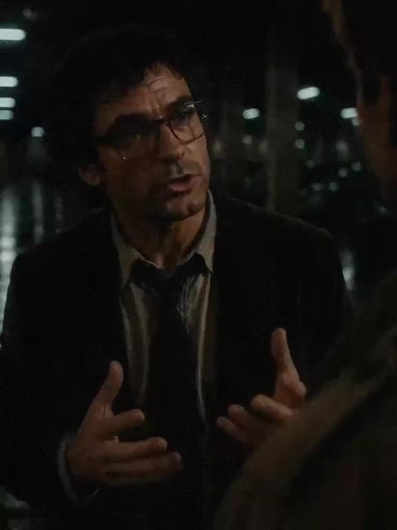 A man in glasses gestures mid-argument under fluorescent light.